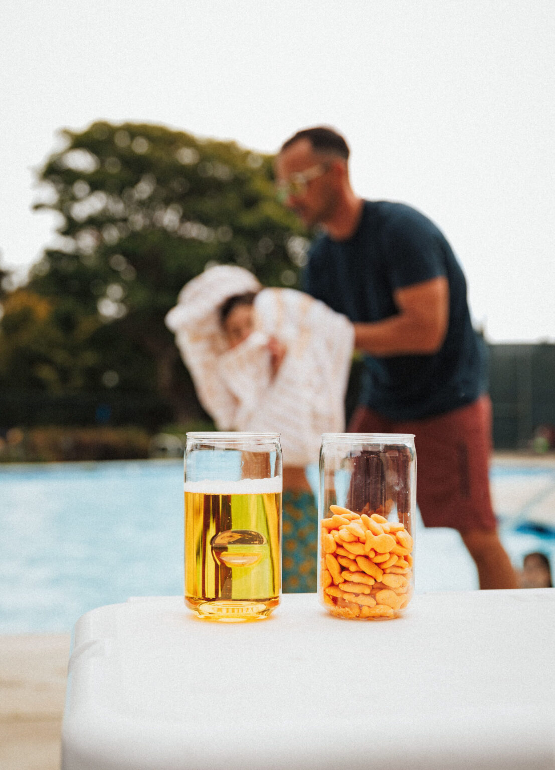 A father drying off his kid at the pool with two govino shatterproof beer can glasses in the foreground outside by a pool